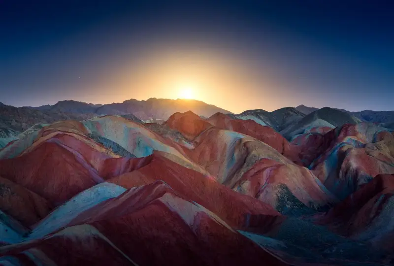 Bunte Schichten aus Rot, Gelb und Blau formen die **Regenbogenberge Chinas** unter einem tiefblauen Himmel mit aufgehender Sonne.