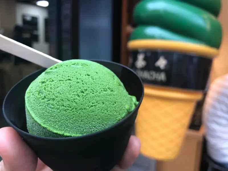 Zielone, japońskie lody matcha w czarnym kubeczku, z drewnianą łyżeczką. W tle wielki, żółty rożek z lodami.