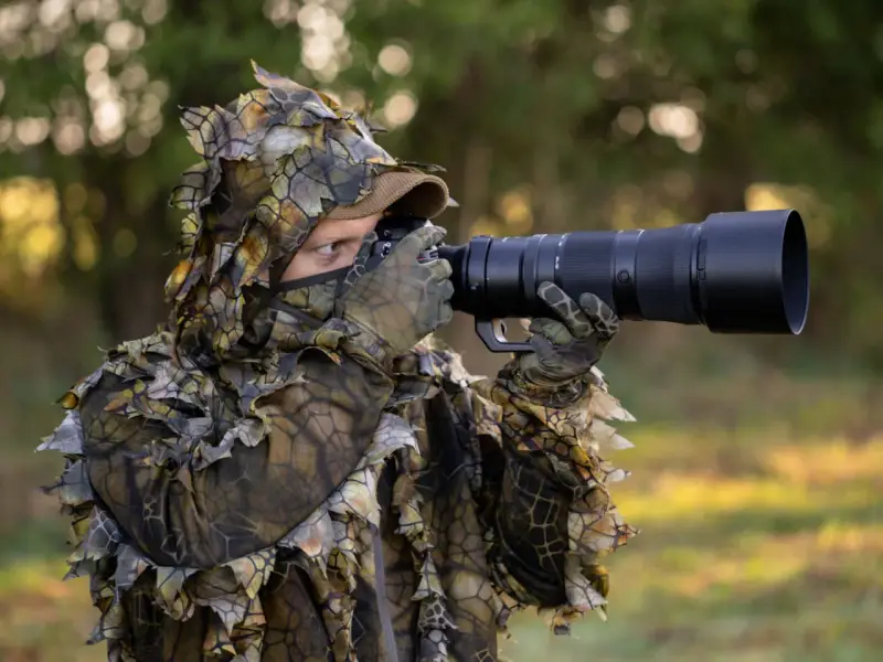 Fotograf w ghillie suit z aparatem do fotografowania zwierząt czeka na idealny kadr.