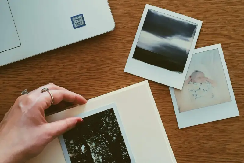 Dłoń układa zdjęcie na albumie, obok inne fotografie i laptop. Idealne miejsce, gdzie przechowywać zdjęcia z telefonu.