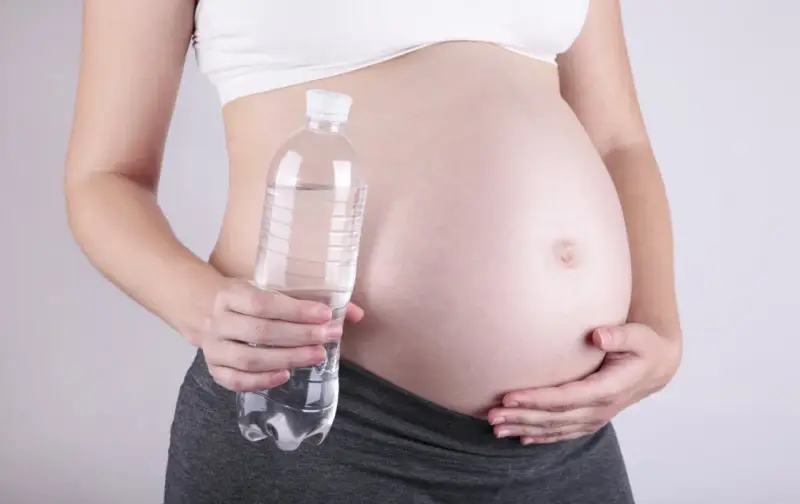 Ciężarna kobieta trzyma butelkę wody, symbolizującą, jak wyglądają wody płodowe – czyste i przejrzyste.