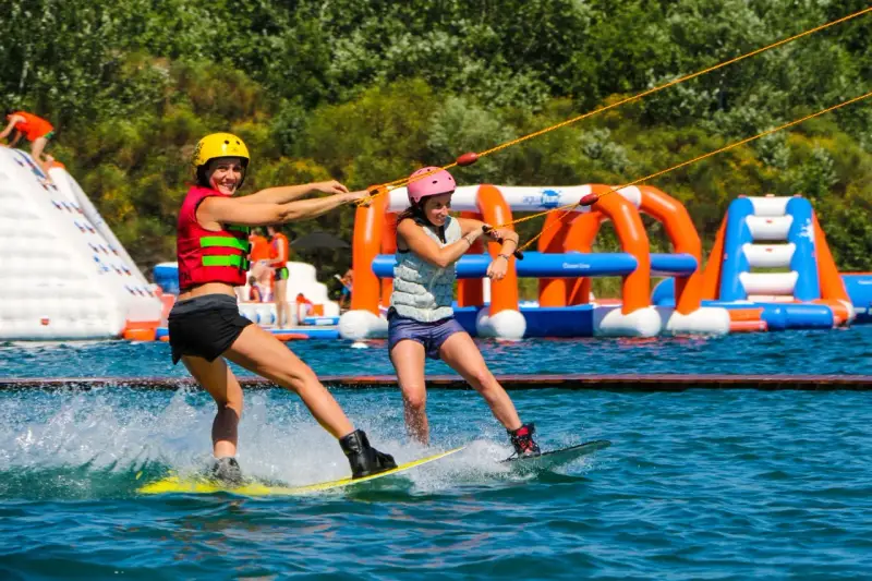 Deux femmes font du wakeboard dans un wam park orange et bleu, avec des structures gonflables en arrière-plan.