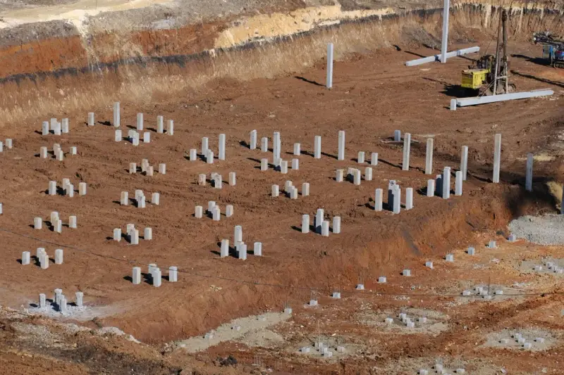 Plac budowy z licznymi betonowymi słupami, tworzącymi fundament pośredni. W tle maszyna budowlana.