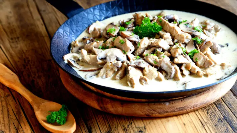 Putengeschnetzeltes mit Pilzen in cremiger Soße, garniert mit Petersilie, serviert in einer Pfanne auf rustikalem Holztisch.