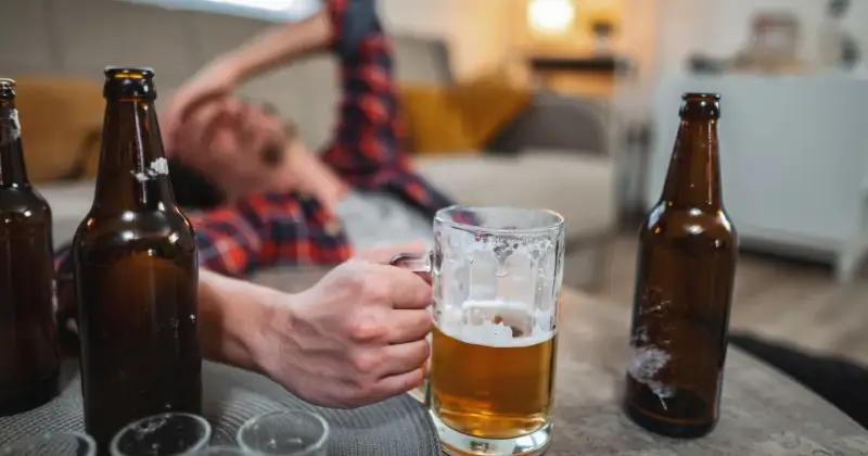 Mężczyzna leży na kanapie, trzymając kufel piwa. Wokół leżą puste butelki. Czy to oznacza, co przyspiesza spalanie alkoholu?