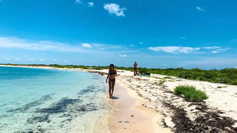 Kobieta spaceruje po piaszczystej plaży nad turkusową wodą. W tle widać innych ludzi i bujną zieleń. To idealne miejsce na wypoczynek nad Zatoką Meksykańską.