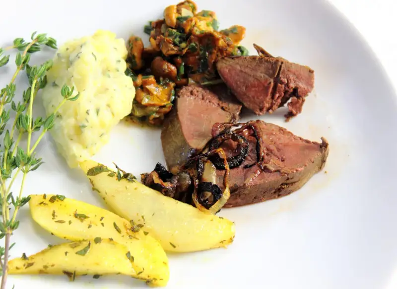 Rehleber zubereiten: zarte Scheiben Rehleber mit gebratenen Zwiebeln, Kartoffelpüree, gebratenen Kartoffelspalten und frischen Kräutern auf einem weißen Teller.