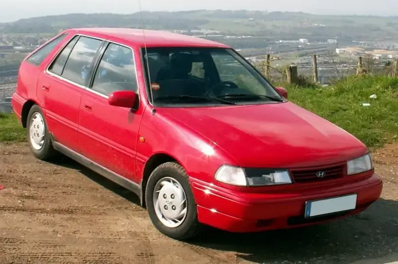 Czerwony Hyundai Pony z lat 90. stoi na polnej drodze, z widokiem na dolinę i miasto w tle.