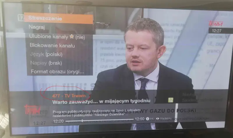 Mężczyzna w garniturze na ekranie telewizora, na którym widać menu z opcją "477 - TV Trwam".