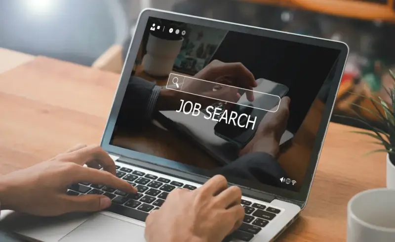 Ręce wpisują "JOB SEARCH" na laptopie. Praca wre, szukamy nowych możliwości.