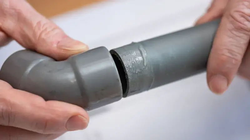Mains de personne assemblant deux tuyaux gris en **pvc renforcé**, l'un avec un coude, prêts pour une installation solide.