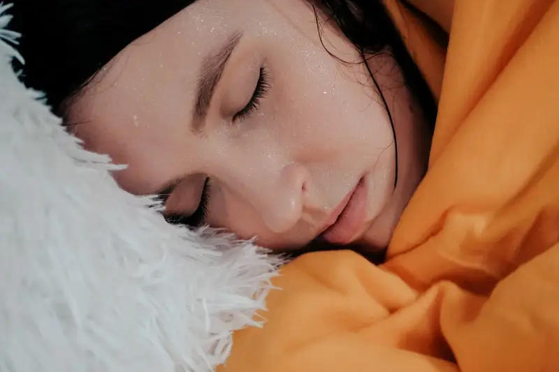 Viso di donna con gocce di sudore sulla fronte, avvolta in una coperta arancione e piumino bianco. Sembra che stia per sudare freddo.