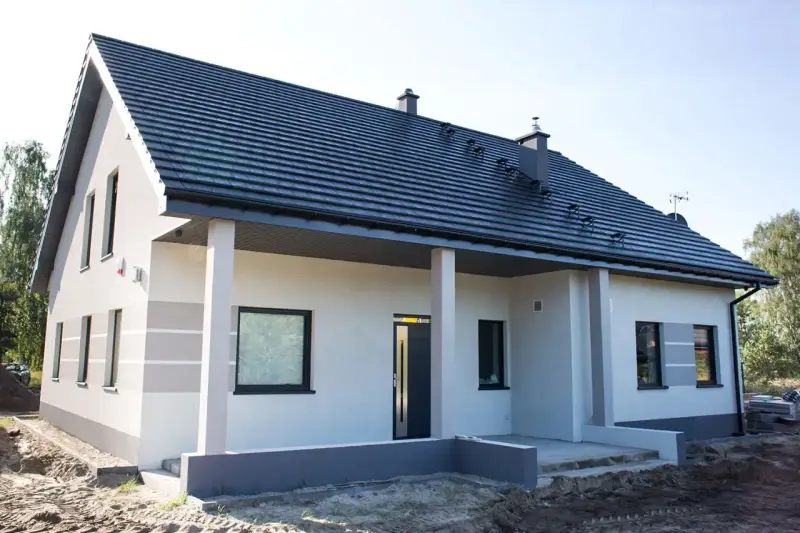Nowy dom z szarą dachówką i jasną elewacją, ozdobioną subtelnymi, poziomymi pasami. Trwają prace przy malowaniu elewacji, tworząc nowoczesny wygląd.