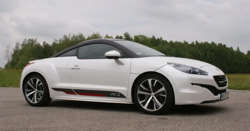 Biały Peugeot RCZ z czarnym dachem i sportowymi akcentami na progu, zaparkowany na tle zielonego lasu.