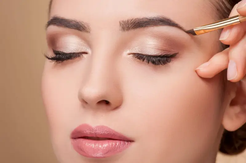Une main applique un maquillage subtil pour parfaire la forme sourcil d'une femme aux yeux fermés.