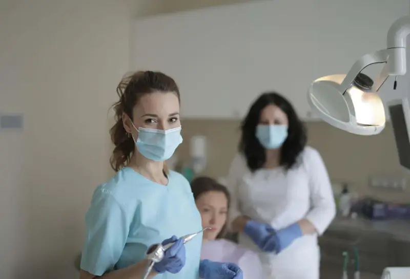 Uśmiechnięta studentka stomatologii w masce i rękawiczkach trzyma wiertło dentystyczne. W tle inne osoby w gabinecie.