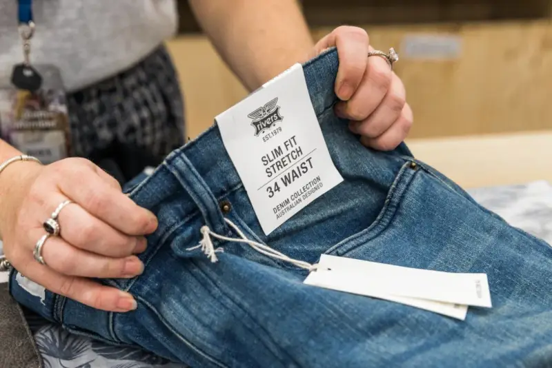 Dłonie trzymają niebieskie jeansy z metką "SLIM FIT STRETCH 34 WAIST". Idealny rozmiar jeansów dla Ciebie.