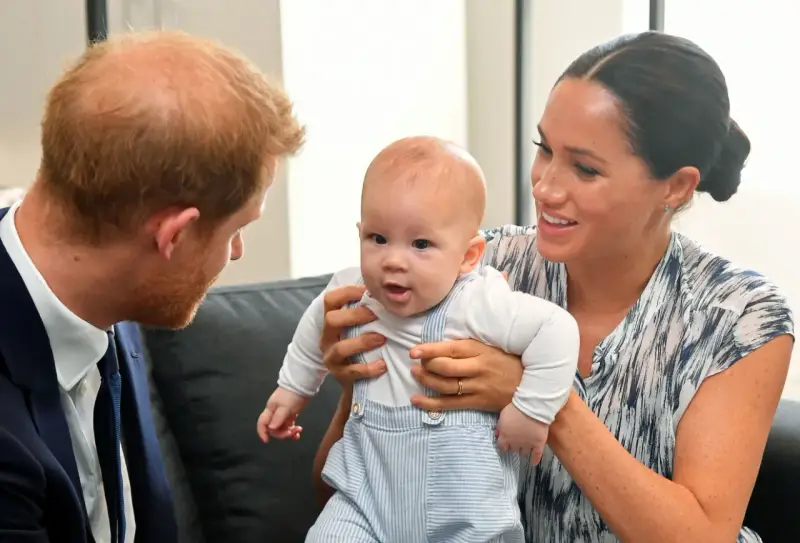 Meghan Markle håller sitt barn, som tittar nyfiket framåt. Prins Harry ser på dem med ett leende.