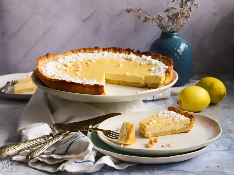 Ein köstliches Zitronentarte Rezept: Eine ganze Tarte und ein Stück auf einem Teller, bestäubt mit Puderzucker. Zwei Zitronen liegen daneben.