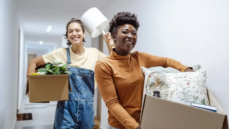 Deux amies souriantes déménagent, portant des cartons remplis d'objets. Une nouvelle aventure commence, peut-être grâce à un **bail solidaire**.