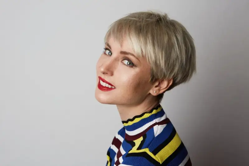Kobieta z blond włosami i grzywką baby bangs, w kolorowym golfie, uśmiecha się do aparatu.