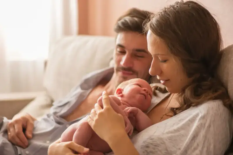 Mama tuli noworodka, tata patrzy z miłością. To piękny obraz opieki na żone po porodzie, pełen czułości i wsparcia.