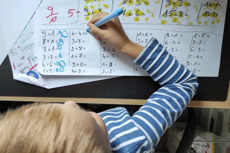 Dziecko rozwiązuje zadania matematyczne. Na kartce widać różne działania, a obok nich cyfry, które mogą stanowić **skalę ocen** lub odpowiedzi.