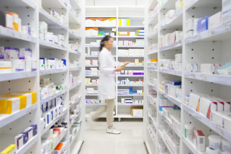 Farmaceutka w białym fartuchu idzie między regałami pełnymi leków, szukając amoksycyliny.