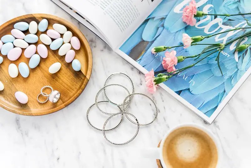 Uroczy flat lay: pastelowe drażetki, srebrna biżuteria, różowe goździki i kawa na marmurowym blacie.