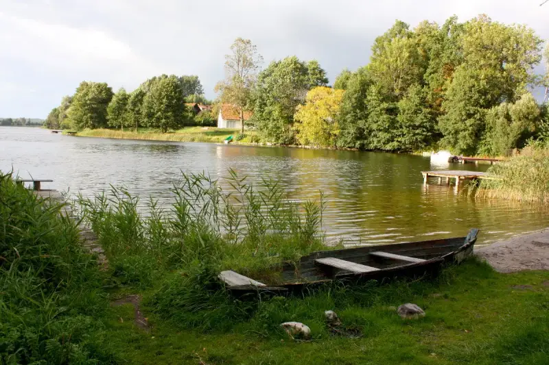 Łódka na brzegu, w tle zielone drzewa i domki nad wodą. Widok na największe jezioro w Polsce.