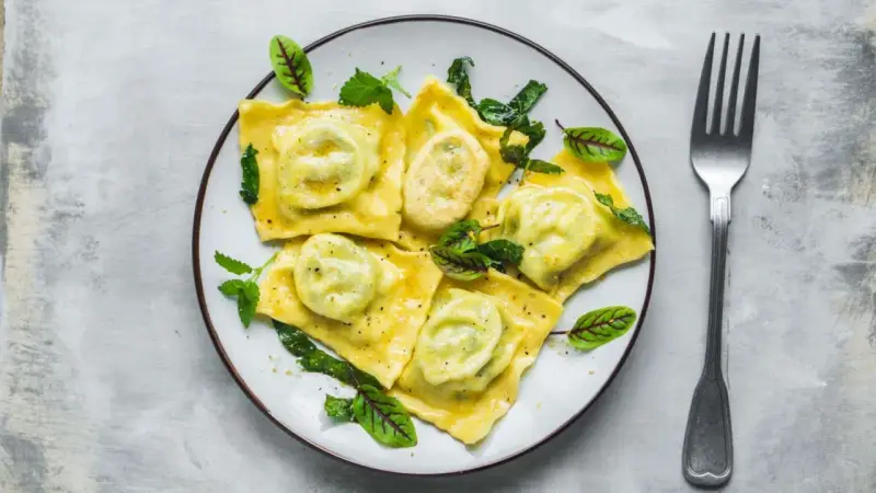 Pyszne ravioli z ricottą i szpinakiem, polane sosem, ozdobione świeżymi ziołami. Obok leży widelec.