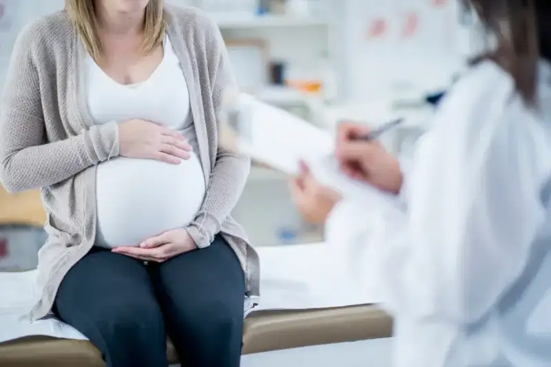 Kobieta w ciąży siedzi na kozetce, trzymając dłonie na brzuchu. Lekarz spisuje dokumenty, prawdopodobnie wystawiając zaświadczenie o ciąży.