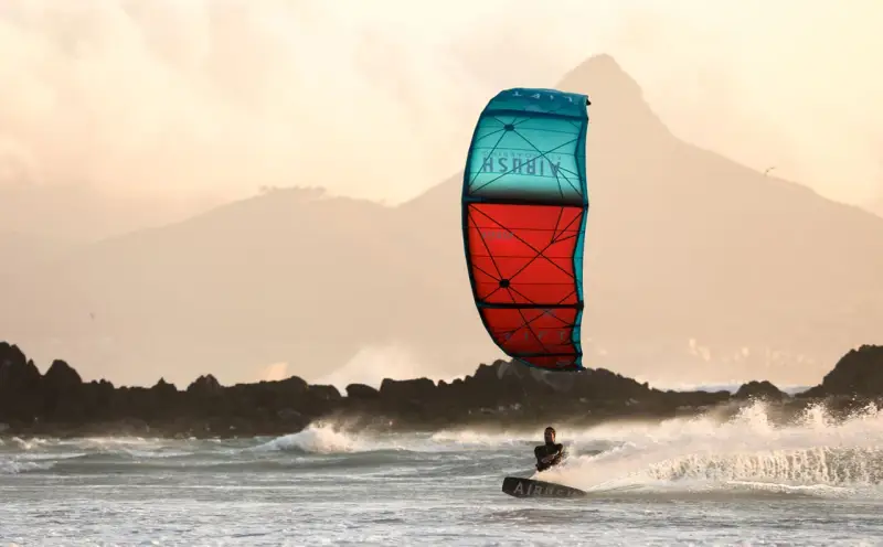 Kitesurfer na desce w akcji, z kolorowym latawcem na tle gór i morza.