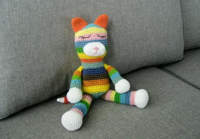 Ett färgglatt virkat gosedjur i form av en katt sitter på en grå soffa.