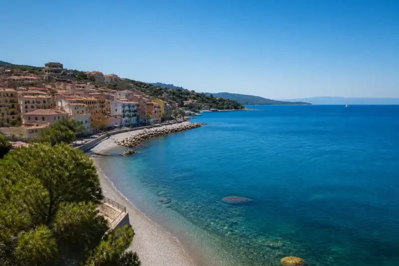 Porto Santo Stefano: case colorate affacciate su spiagge di ciottoli e sabbia, con il mare azzurro e un veliero all'orizzonte.