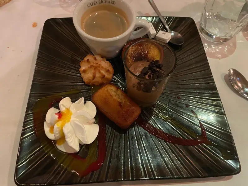 Un assortiment gourmand : café, financier, macaron, verrine chocolat et chantilly, servi sur une assiette noire texturée, digne de la taverne des fougères.