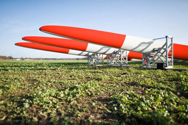 Drei Rotorblätter für Windkraftanlagen liegen auf einem Feld. Sie sind rot-weiß gestreift und warten auf ihren Einsatz nach dem EEG.