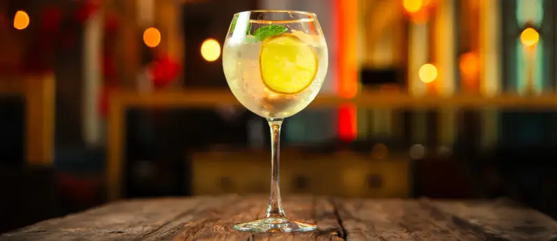 Erfrischender Martini Bianco Spritz mit Zitrone und Minze auf einem Holztisch, im Hintergrund unscharfe Lichter.