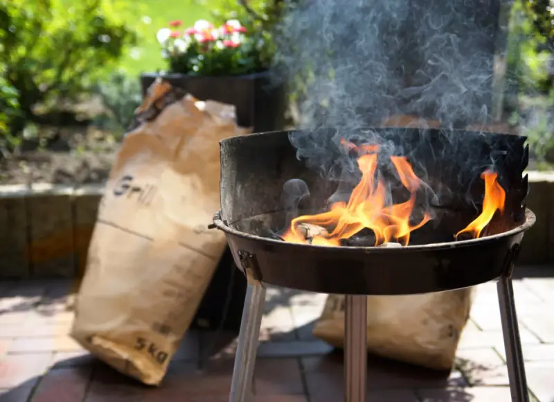 Rozpalony grill z płomieniami i dymem, obok torba z węglem. Idealny początek, by dowiedzieć się, jak rozpalić grilla.