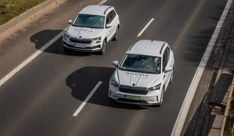 Dwa białe samochody Skoda Auto Szkoła jadą po drodze, gdzie obowiązuje ruch lewostronny.