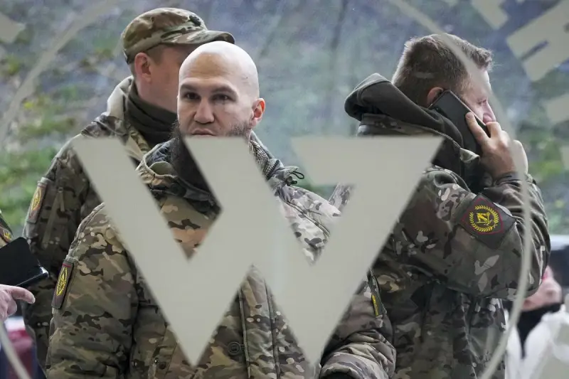 Trzech mężczyzn w mundurach, jeden rozmawia przez telefon, na tle symbolu przypominającego literę "W". Mogą to być członkowie grupy Wagnera.