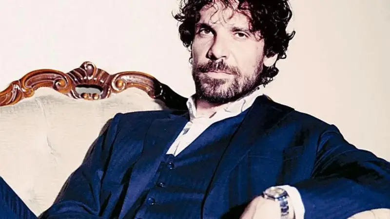 L'attore Montanari, con barba e capelli ricci, indossa un completo blu e una camicia bianca, seduto su una poltrona d'epoca.