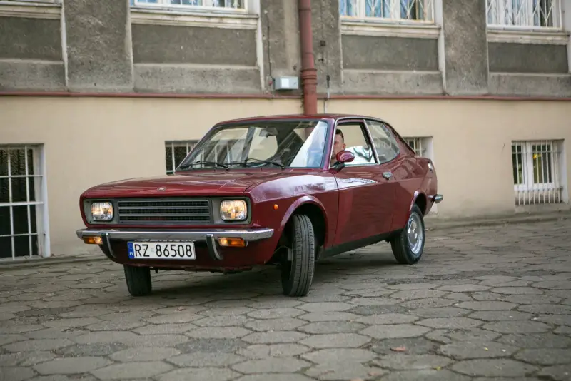 Czerwony fiat 128, klasyk z lat 70., zaparkowany na brukowanym dziedzińcu przed budynkiem.