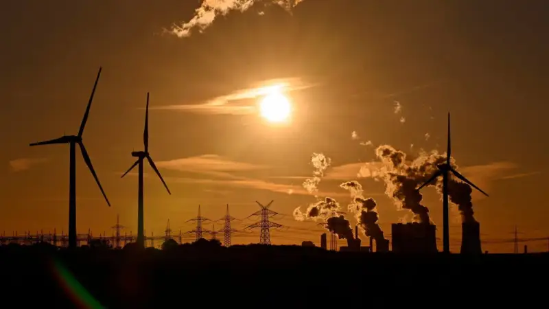 Silhouetten von Windrädern und rauchenden Industrieanlagen im Sonnenuntergang. Ein Bild, das zum Nachdenken über Stromkosten Norwegen vs. Deutschland anregt.