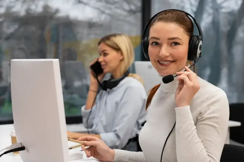Uśmiechnięta key account manager w słuchawkach z mikrofonem pracuje przy komputerze. W tle koleżanka rozmawia przez telefon.