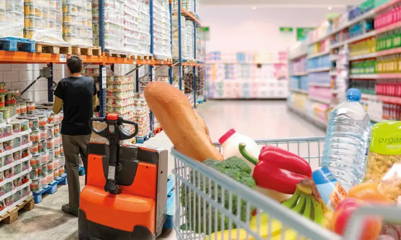 W magazynie FMCG, pracownik obsługuje wózek widłowy, podczas gdy wózek sklepowy pełen produktów czeka na zakupy.
