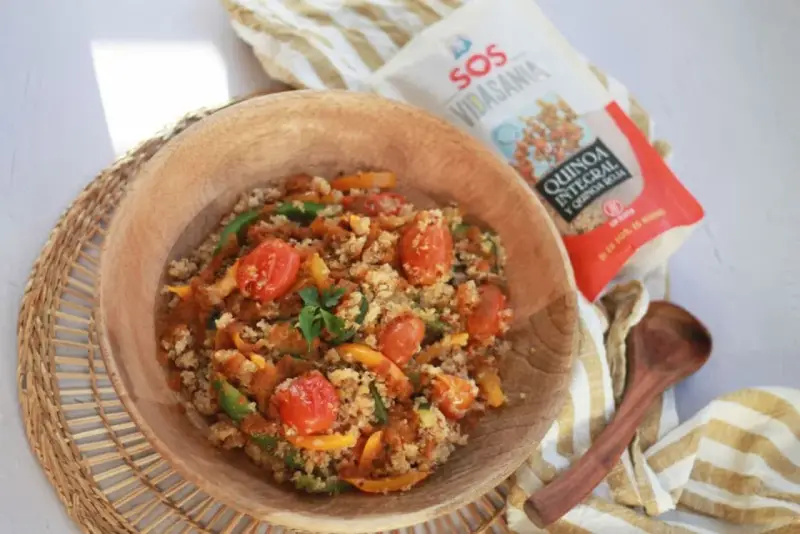 Un tazón de madera lleno de quinoa con verduras coloridas: tomates cherry, pimientos verdes y amarillos. Cerca, un paquete de quinoa integral y roja.