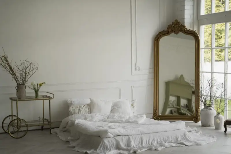 Elegancka sypialnia z łóżkiem w bieli, ozdobiona sztukaterią przypominającą lamperię angielską. Złote lustro i wózek dodają blasku.