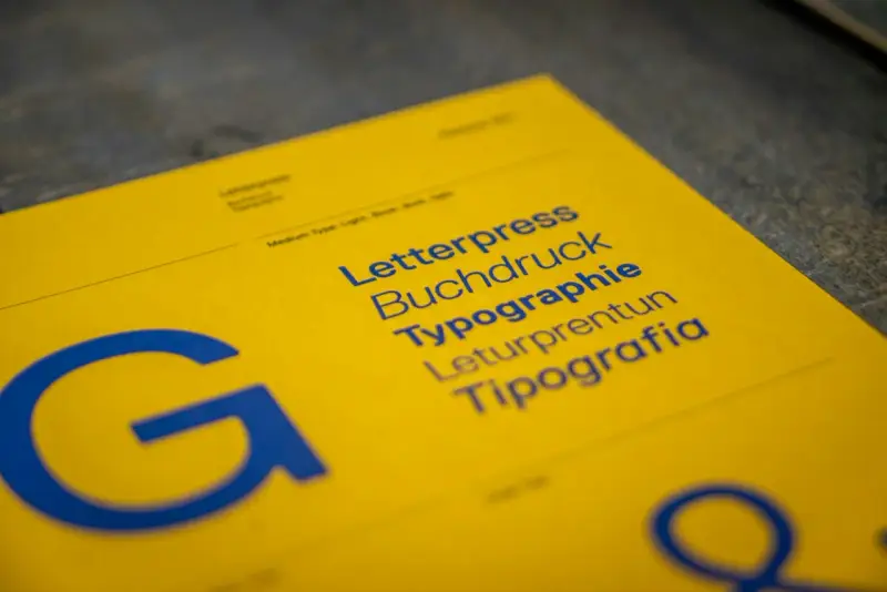 Żółta kartka z niebieskimi napisami: Letterpress, Buchdruck, Typographie, Leturpretun, Tipografia. Duża litera "G" po lewej. To praca typografa.