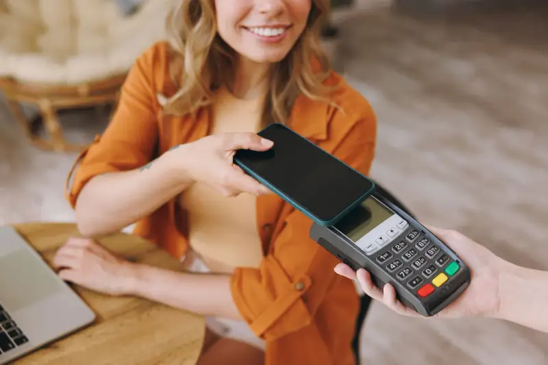Uśmiechnięta kobieta zbliża telefon do terminala płatniczego, pokazując, jak płacić telefonem. Szybko i wygodnie.